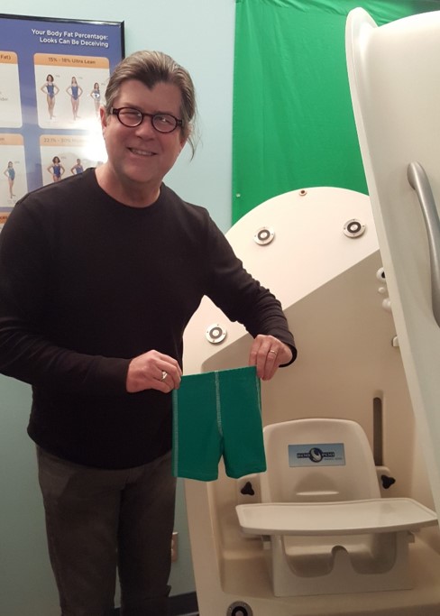 Dr. Shepherd holding the small green compression shorts of the Tod Pod Pediatric Option, next to the Bod Pod chamber